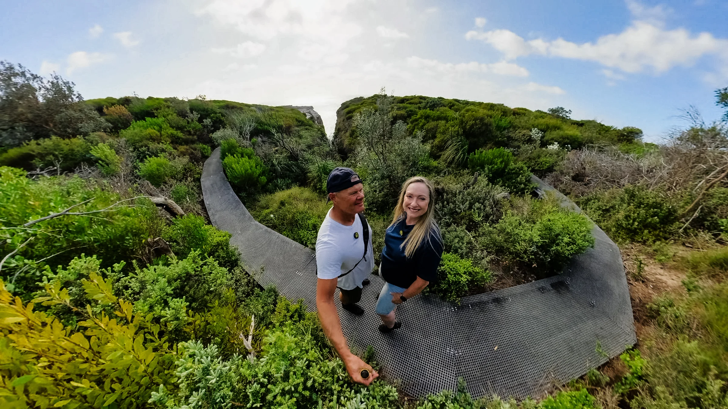 Rob and Sue smiling on a curved boardwalk among coastal greenery
