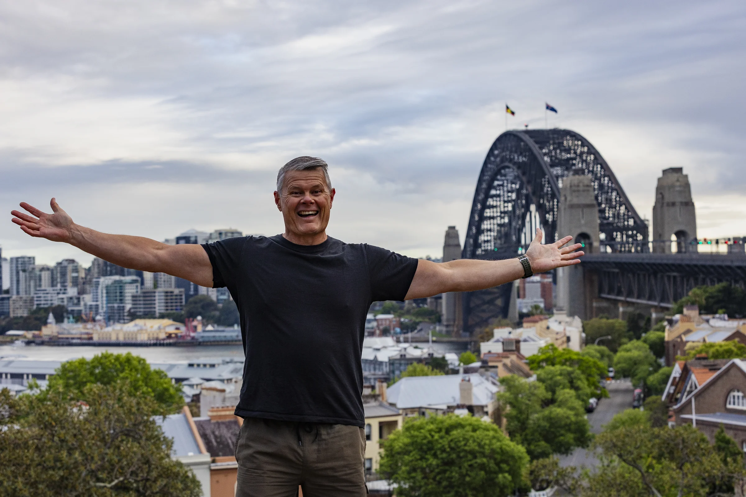 Rob smiling with his arms outstretched and the Harbour Bridge beyond Observatory Hill
