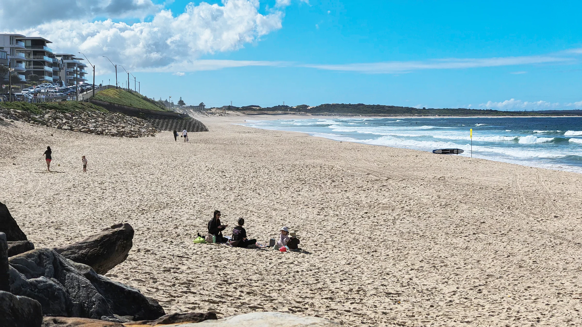 People sitting on North Cronulla Beach with the promenade, sand and breaking waves beyond