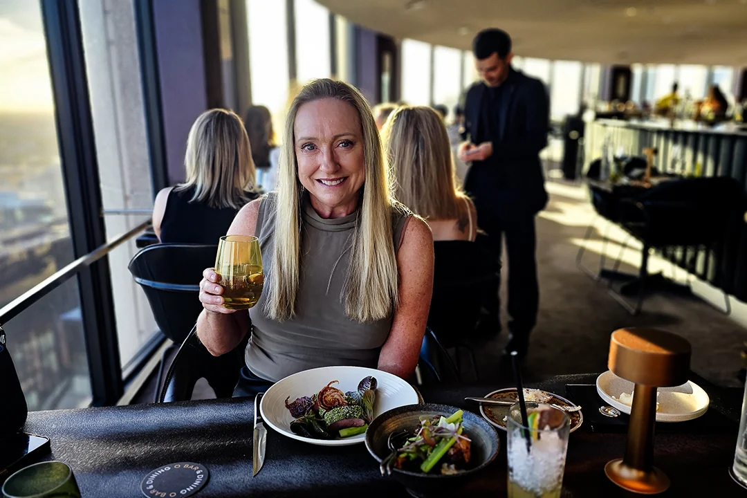 Sue enjoying a meal and drink at O Bar, with the restaurant and city view behind her